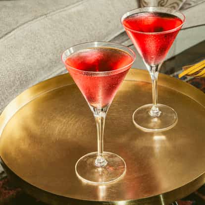Pink Martini cocktails in chilled martini glasses on a gold tray beside a sofa and colorful books on a glass coffee table.
