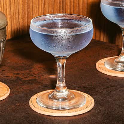 Purple Cosmopolitan in gekühlten Coupegläsern mit beschlagenen Rändern, serviert auf Lederuntersetzern auf einer dunklen Bartheke.