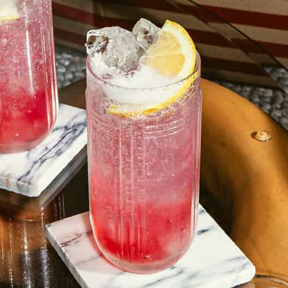 Red Tonic cocktail in tall ribbed glass with ice and lemon wedge, sparkling red drink on marble coaster atop a glass table.