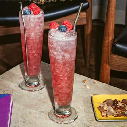 Koktajl Russian Spring Punch w wysokiej szklance z kruszonym lodem, różowym kolorem, udekorowany malinami i jagodą, ustawiony na kamiennym stole.