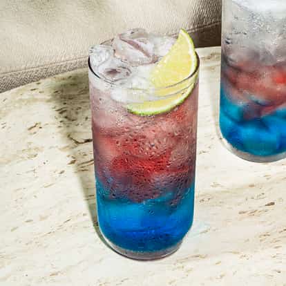 Koktajl Sparkler w wysokiej szklance z warstwami niebieskiego i czerwonego koloru, z lodem i limonką.