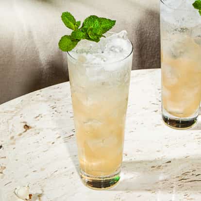 Drink Swedish Death Nettle em copo alto com gelo e hortelã fresca, bebida dourada clara sobre mesa de pedra.