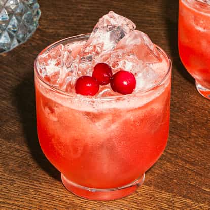 Ein Sweetheart Cocktail im Tumblerglas mit Eiswürfeln und roten Cranberrys, ein leuchtend rosaroter Drink auf einem Holztisch.