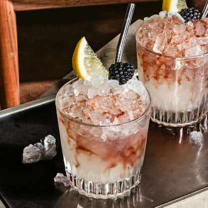 Vodka Bramble Cocktail im Tumblerglas mit Crushed Ice, rosafarbenen Wirbeln und Garnitur aus Zitrone und Brombeere, auf einem Metalltablett serviert.