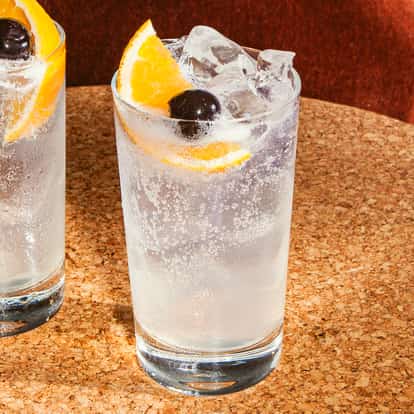 Vodka-Collins-Cocktail im hohen Glas mit klarer Kohlensäure, Eiswürfeln sowie einer Orangenscheibe und dunkler Kirsche auf einer Korkoberfläche.