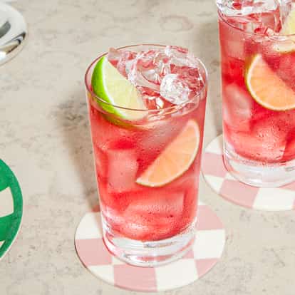 Two vodka cranberry cocktails with ice and lime wedges on patterned coasters beside a plate of fresh limes on a light stone surface.