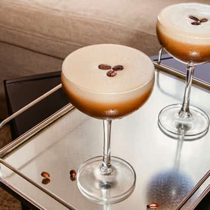 Vodka Espresso cocktails in coupe glasses topped with coffee beans on a mirrored tray beside a sofa.