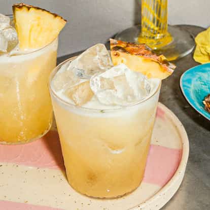 Vodka Fizz cocktails in ice-filled glasses with pineapple garnishes on a pink-striped plate beside a dish of fresh pineapple slices.