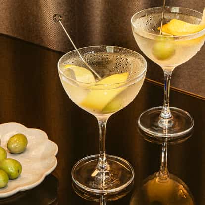 Two vodka martini cocktails with lemon peel and green olives on a dark glossy table beside a plate of fresh green olives.
