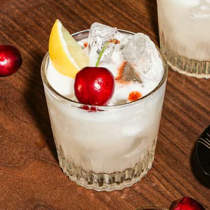Cóctel vodka sour con hielo, rodaja de limón, cereza y bitter, servido en vaso corto sobre bandeja de madera.