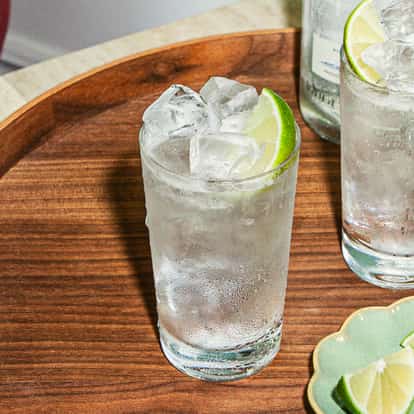 Dwa koktajle wódka tonic z lodem i limonką na drewnianej tacy obok małego talerzyka z kawałkami limonki.