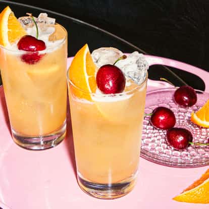 Yellowhammer w wysokich szklankach z lodem, z pomarańczami i czereśniami na różowej tacy obok talerzyka z czereśniami i kawałkami pomarańczy.