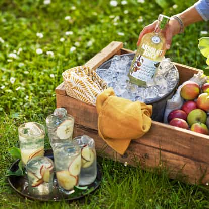 Absolut Juice Apple bottle in an ice-filled metal bucket with apples and four glasses on a tray on grass