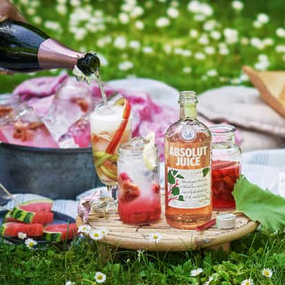 Absolut Juice Rhubarb bottle on a round wooden board with two jars of rhubarb and ice and a tall glass being filled