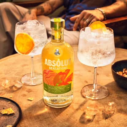 Absolut Sensations bottle on wooden table with two stemmed glasses filled with ice and orange slice garnishes