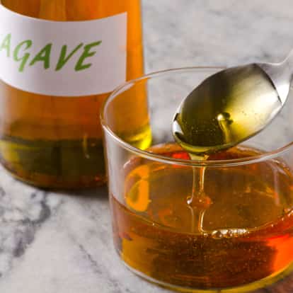 Agave Nectar in a glass bowl with syrup dripping from a spoon and a bottle labeled Agave on marble