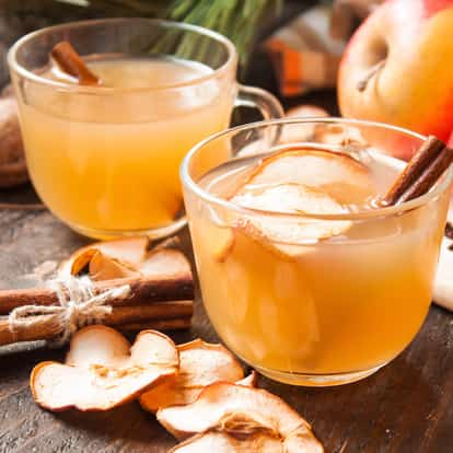 Apple Juice (hot) in two clear glass mugs with apple slices and a cinnamon stick on a wooden surface