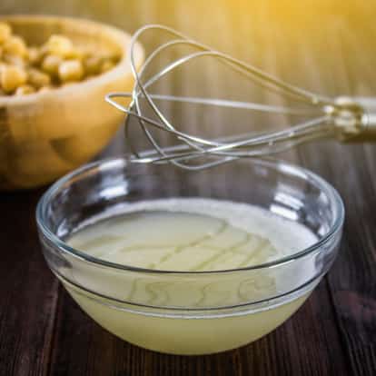 Aquafaba in a glass bowl with a whisk and a bowl of chickpeas on a wooden surface