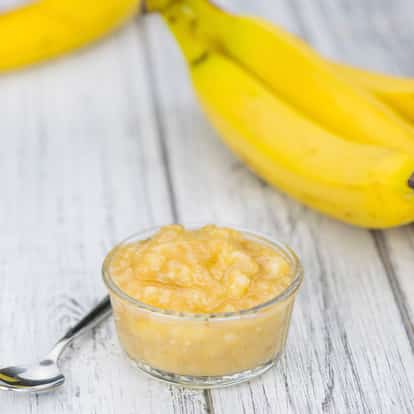 Banana puree in a small glass bowl with a spoon and two bananas on a wooden surface