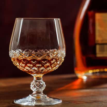 Benedictine Dom in a cut crystal snifter on a wooden surface with a bottle in the background