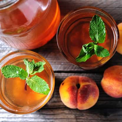 Bourbon, Peach Flavored in two short glasses with mint sprigs and whole peaches on a wooden surface