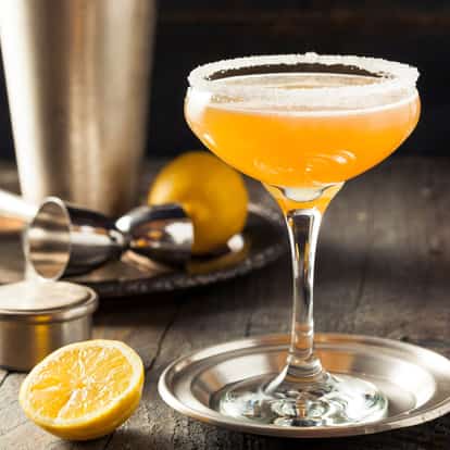 Caperitif served in a sugar-rimmed coupe on a metal coaster with lemon half and bar tools on a wooden surface