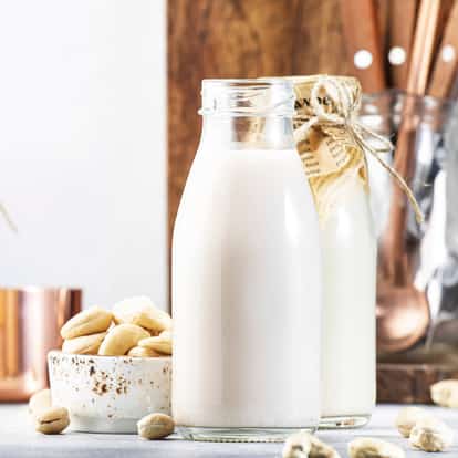 Cashew milk in two glass bottles one open and one wrapped in paper with a bowl of cashews on white surface