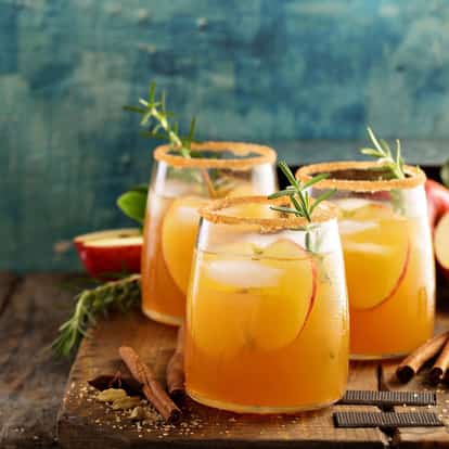 Cider in three rocks glasses with ice, apple slices, rosemary sprigs and brown sugar rims on a wooden board