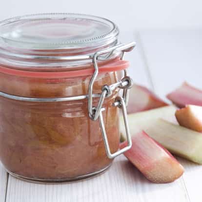 Cinnamon Spiced Rhubarb Puree in a glass jar with metal clasp beside rhubarb pieces on a white wooden surface