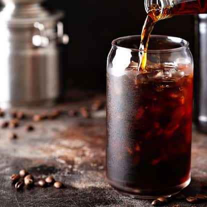 Cold Brew Coffee in a tall glass with ice being poured from a bottle on a dark tabletop