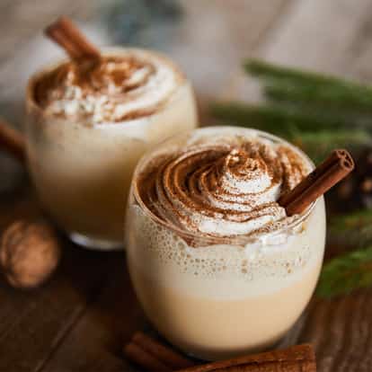 Eggnog in two stemless glasses topped with whipped cream sprinkled with ground cinnamon and cinnamon sticks
