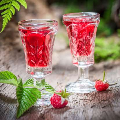 Fresh Homemade Raspberry Cordial served in two etched cordial glasses with raspberries and leaves on wooden surface