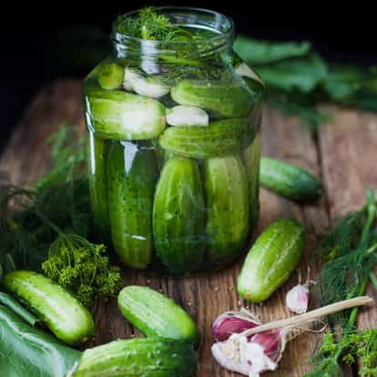A jar of pickled gherkins with garlic cloves and dill on a wooden board.