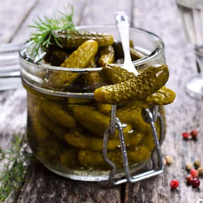 Gherkin pickles sit in a glass clamp jar with a metal fork lifting one on a wooden table.