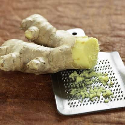 Ginger (grated) on a metal grater with an unpeeled ginger root on a textured background