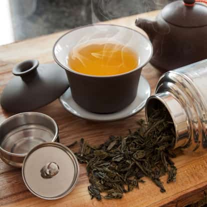 Hot green oolong tea in a gaiwan with loose leaves and a teapot on a wooden board.