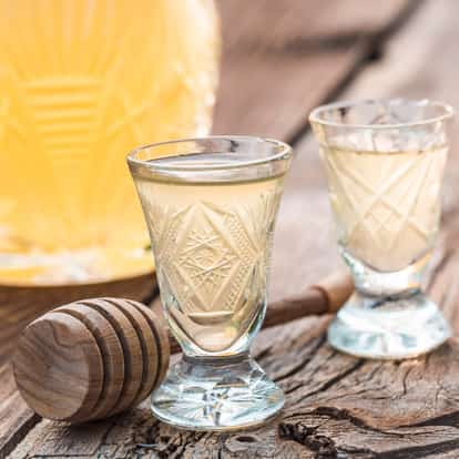 Two cut-glass cordial glasses filled with pale honey liqueur beside a wooden honey dipper on rustic wood.