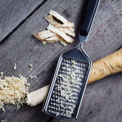 Horseradish root with peeled strips and grated shavings beside a stainless steel grater on a wooden board