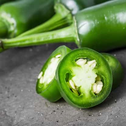 Jalapeno sliced into rounds with seeds visible and whole peppers in the background on a stone surface