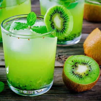 Kiwi Liqueur in a rocks glass with ice and mint sprig, kiwi wheel on the rim and halved kiwi on wood