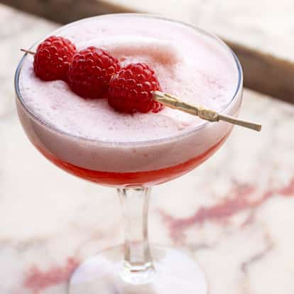 Lillet Rouge in a coupe glass with foam and three raspberries on a wooden pick on a marble surface