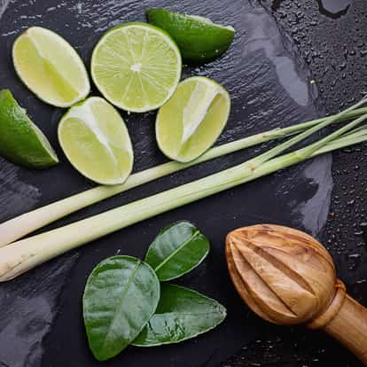Lime and lemongrass cordial halved limes lemongrass stalks kaffir lime leaves and wooden reamer on black slate