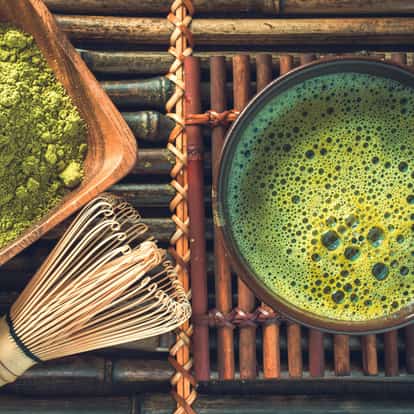Matcha in a bowl with froth, powder in a wooden dish and a bamboo whisk on a woven mat