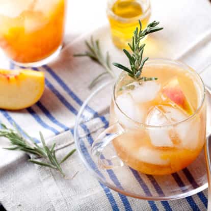 Peach Liqueur in a glass mug with ice, peach wedge and rosemary sprig on striped cloth