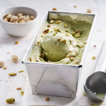 Pistachio ice cream in a metal loaf pan with a scoop and scattered pistachio nuts on white background