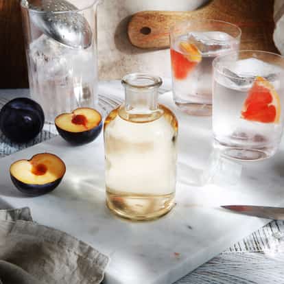 Plum Wine in a small glass bottle with halved plums and two rocks glasses with ice cubes and citrus wedges