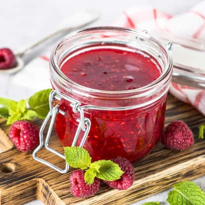 Raspberry jam in a clamp-lid glass jar on a wooden board with raspberries and mint sprigs