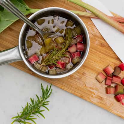 Rhubarb and rosemary syrup in a small saucepan with diced rhubarb and a rosemary sprig on a wooden cutting board