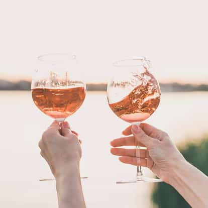 Two wine glasses of rosé sparkling wine held aloft by hands at a lake at sunset.