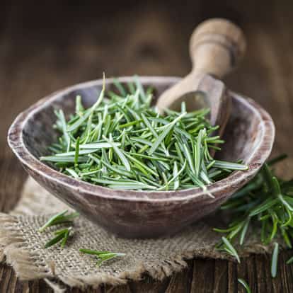 Rosemary sprigs in a wooden bowl with a wooden pestle on burlap atop a wooden table